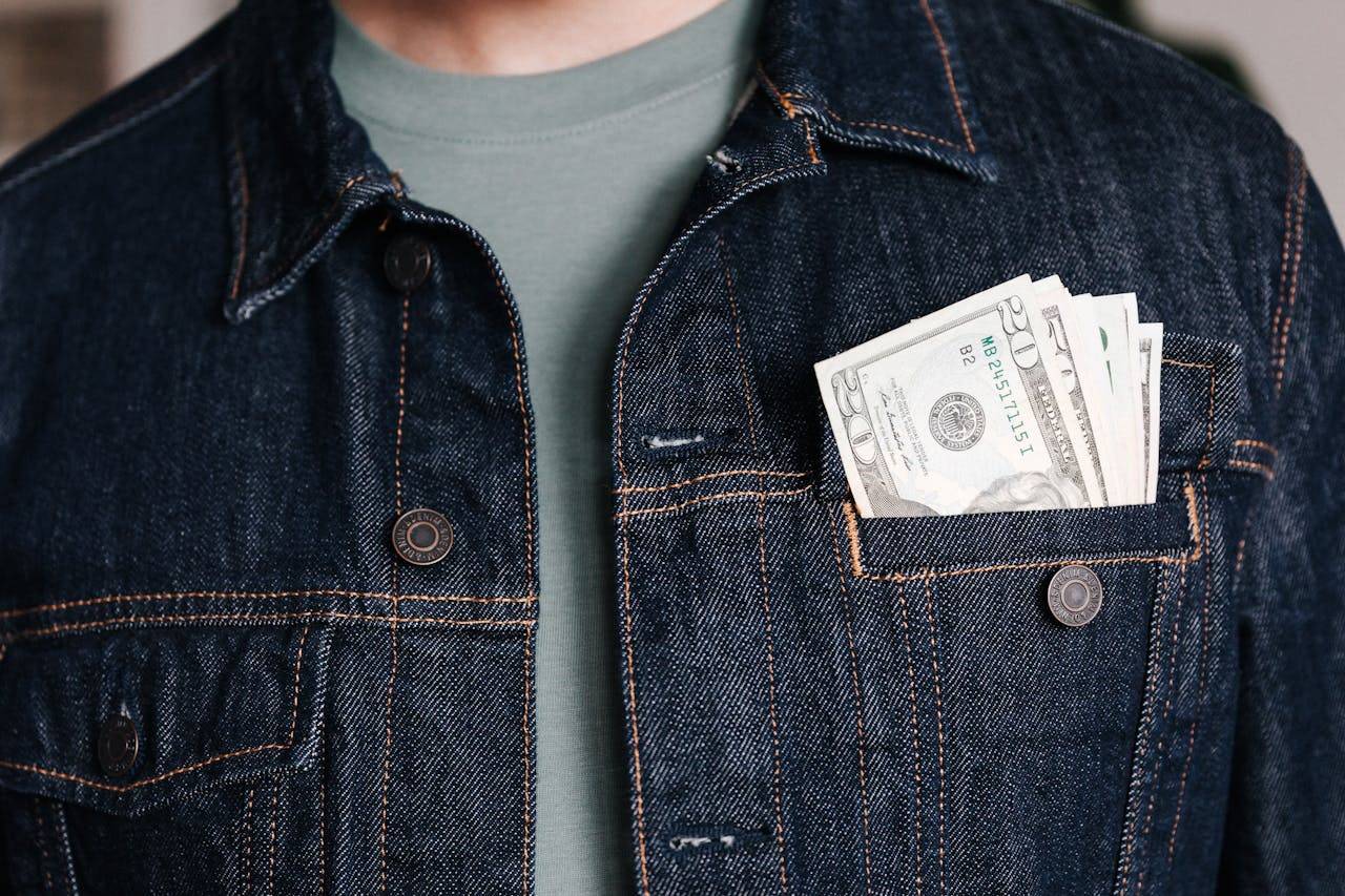 Crop unrecognizable male in casual outfit standing with different nominal pars of dollar banknotes in pocket of jeans jacket